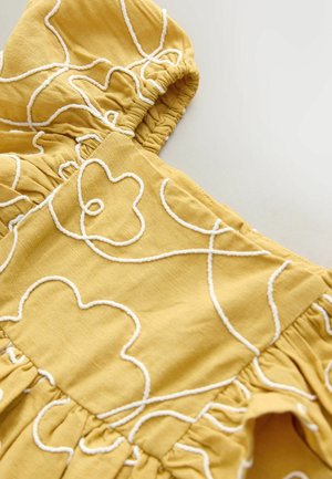 Žlutá látka s bílými vyšívanými květinovými vzory, nabíraným výstřihem, texturovaným povrchem a rozšířenými záhyby na spodní straně.