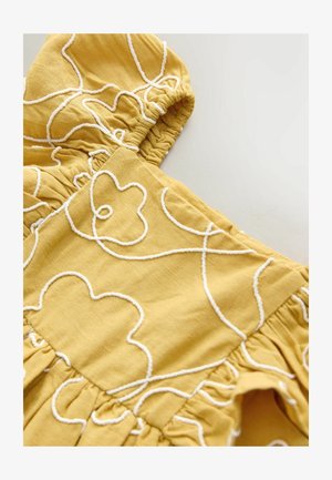 Žlutá látka s bílými vyšívanými květinovými vzory, nabíraným výstřihem, texturovaným povrchem a rozšířenými záhyby na spodní straně.