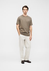 T-shirt en coton marron à manches courtes et col rond, associé à un pantalon ample de couleur claire et des chaussures foncées.