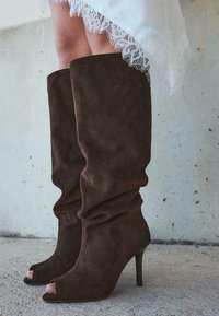 Brown suede knee-high boots with a slouchy design, open-toe style, and a stiletto heel. Textured surface with visible stitching details.