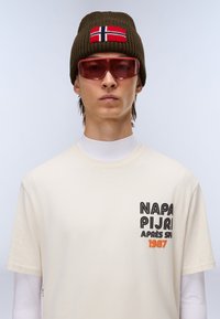 Lys beige t-shirt med rund hals og grafisk tekst i sort og orange. Brun strikket hue med et rødt og hvidt norsk flag. Lyserøde solbriller.