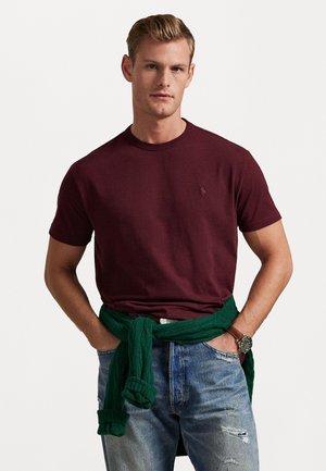 Burgunderfarvet t-shirt med korte ærmer og rund halsudskæring, parret med lyse jeans. Mørkegrøn sweater bundet om livet. Ur på håndleddet.