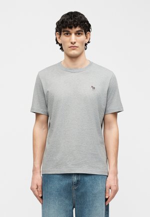 Νεαρός άνδρας με κοντά, σγουρά μαλλιά, που φοράει ένα απλό ανοιχτό γκρι t-shirt με ένα μικρό λογότυπο ζέβρας στο στήθος και μπλε τζιν, στέκεται αντικρίζοντας ευθεία μπροστά.