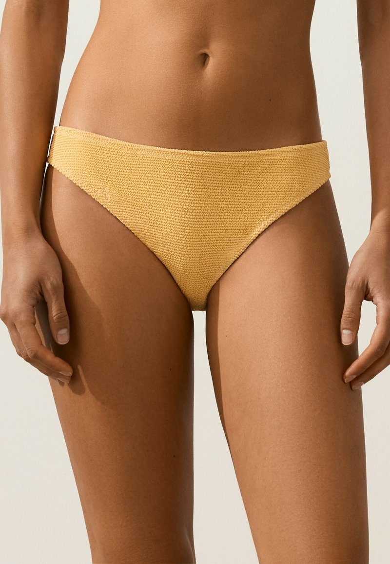 Gros plan du bas du torse portant un bas de bikini jaune texturé avec les mains détendues sur les côtés sur un fond clair.