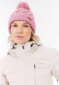 Roze gebreide muts met pompon-detail; gecombineerd met een lichtgekleurde geïsoleerde jas met een rits en een logo op de borst.
