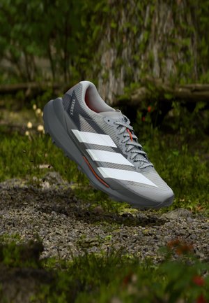 Scarpa da corsa grigia con strisce bianche, tomaia in mesh, dettagli arancioni e suola in gomma traforata, sospesa sopra uno sfondo di ghiaia e erba.