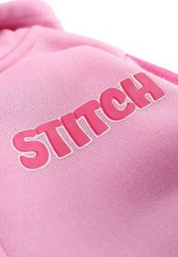 Sweat à capuche rose avec "STITCH" imprimé en gros caractères roses. Le tissu a une texture douce avec des détails de couture visibles.