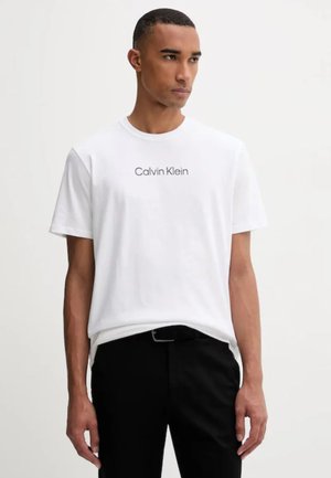 Uomo che indossa una t-shirt bianca Calvin Klein e pantaloni neri, in piedi davanti a uno sfondo grigio chiaro uniforme, guardando di lato.