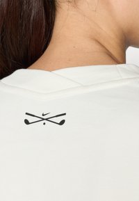 Λευκό πουκάμισο πόλο απεικονιζόμενο από πίσω, με το μαύρο λογότυπο Nike και διασταυρωμένα μπαστούνια γκολφ στον λαιμό. Υφή υφάσματος και δομημένος γιακάς.