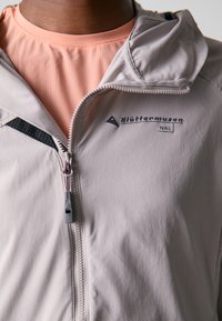 Giacca zip grigia chiara in tessuto testurizzato, con un accento nero, cappuccio e etichetta con logo. Indossata sopra una maglietta rosa a maniche corte.