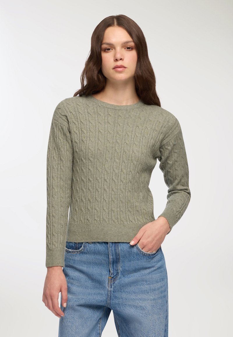 Kobieta z długimi brązowymi włosami, ubrana w jasnoszary sweter w warkocze i niebieskie dżinsy, stojąca z jedną ręką w kieszeni na białym tle.