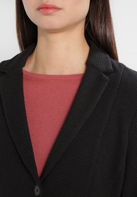 Czarny żakiet o fakturowanej powierzchni z klapami, noszony na różowej bluzce. Żakiet ma zapięcie na jeden guzik i zachowuje strukturalny kroj.