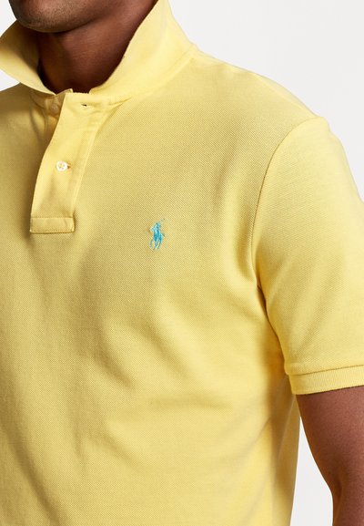 Camisa polo amarela feita de tecido texturizado, com colarinho e um logo azul bordado discreto no lado esquerdo do peito. Duas botões no pescoço.