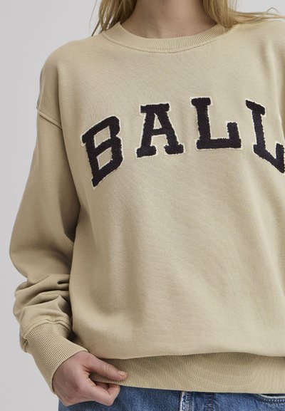 Beige sweatshirt med svart frotté skrift "BALL." Den har rund halslinning og ribbestrikkede mansjetter. Modellen holder i underkanten.
