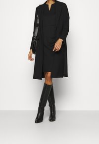 Musta, vartalonmyötäinen mekko V-pouvalla, yhdistettynä pitkään, avoimeen mustaan takkiin. Mallilla on polveen saakka yltävät mustat nahkasaappaat ja hän pitää kädessään mustaa laukkua.