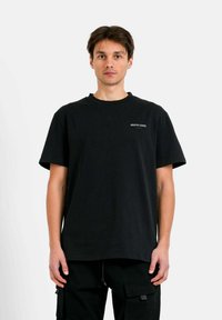 Zwarte katoenen t-shirt met korte mouwen, ronde hals en een klein wit logo op de linkerborst. De pasvorm is relaxed en iets oversized.