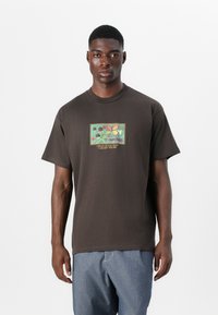 T-shirt en coton marron avec un imprimé graphique rectangulaire et coloré représentant des feuilles et des fleurs, centré sur le devant. Manches courtes et col rond.