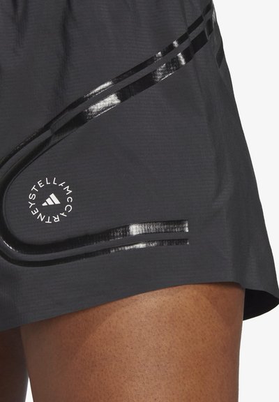 Pantaloni corti sportivi grigi con una texture liscia, accenti neri riflettenti e un logo circolare sull'anca. Le cuciture visibili migliorano il design.