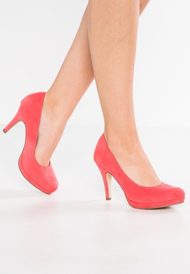 Jambes portant des escarpins à talons hauts en daim rose corail, un pied légèrement levé, sur un fond blanc uni.