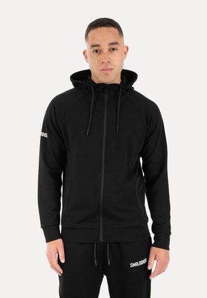 Mand iført sort lynlåshoodie og matchende sorte joggingbukser med hvid "SMILODOX" tekst på bukser og ærme, stående mod hvid baggrund.