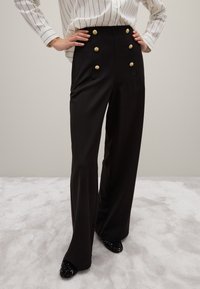 Zwarte hooggewaide broek met accenten van gouden knopen, gemaakt van een gladde stof, met een wijde pijp en een getailleerde silhoeet.