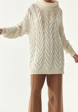 Pull oversized en maille torsadée crème avec un col roulé haut et côtelé. Manches longues et texturées. Associé à un pantalon évasé marron.