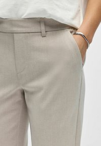 Personne portant un pantalon beige sur mesure, main dans la poche et un bracelet argenté au poignet, associé à un haut blanc.