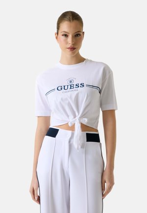 Jeune femme portant un crop top blanc noué avec le texte « Guess California » et un pantalon taille haute blanc avec des accents noirs, debout devant un fond uni.