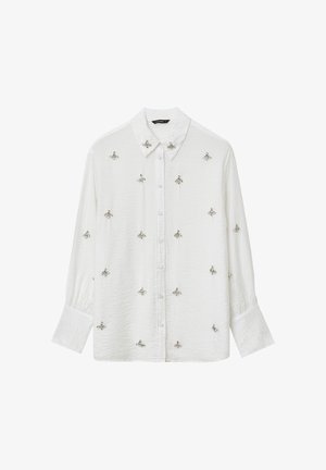 Chemise blanche à boutons avec un tissu texturé, ornée de broderies d'abeilles en argent sur le devant et de manches longues légèrement bouffantes.