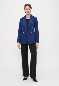 Tummansininen kaksirivinen bleiseri kultanapun kanssa, yhdistettynä mustiin teksturoituihin housuihin, seisoo korkokengissä yksinkertaista taustaa vasten.