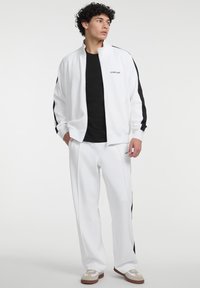 Weißer Zip-Jacket mit schwarzen Seitenstreifen, kombiniert mit einem schwarzen T-Shirt und weißen Jogginghosen. Die Sneaker haben beige und braune Akzente.