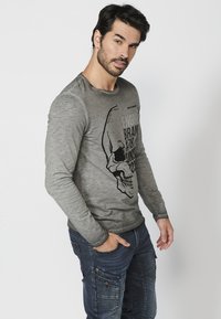 Pilkos spalvos ilgam rankovių marškiniai su juodu kaukolių grafiku ir užrašų spauda. Dėvimi su mėlynomis kargo kelnėmis, kuriose matomos kišenės ir užtrauktukai.