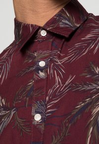 Koszula z krótkim rękawem w bordowym materiale z botanicznym nadrukiem. Detale obejmują złożony kołnierz i białe guziki z przodu.