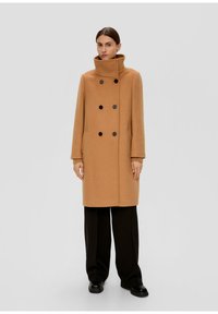 Manteau en laine de chameau avec un col haut, design double boutonnage, boutons noirs et poches latérales. Associé à un pantalon large de couleur sombre.