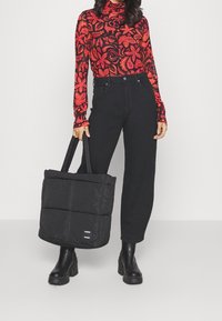 Borsa tote nera imbottita con design a trapunta, accentuata da due manici. Il modello indossa un top a maniche lunghe a fantasia rossa e jeans neri a vita alta.