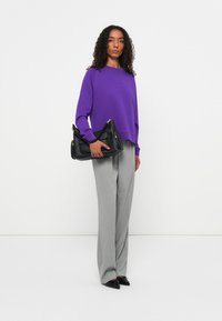 Maglione viola a maniche lunghe abbinato a pantaloni grigi a righe. La modella tiene una borsa strutturata nera con accenti dorati.
