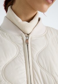 Lys beige quilted bomberjakke med en ribbestrikket kremfarget høyhalset krage, sølvglidelås og teksturerte detaljer i hele stoffet.