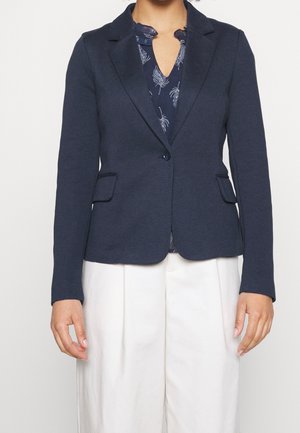 Mujer que lleva una blazer ajustada azul marino con bolsillos con solapa sobre una blusa azul oscuro con estampados claros de plumas y pantalones de cintura alta color crema.