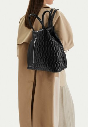 Mujer con abrigo tipo trench beige que lleva una gran mochila de cuero acolchado negra con dos bolsillos verticales con cremallera.