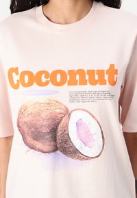 Lichtroze t-shirt met een afbeelding van kokosnoten, met een gedetailleerde bruine schaal en een witte binnenkant. Vet oranje tekst leest "Kokosnoot."
