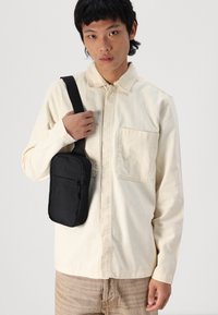 Black crossbody bag with smooth texture, zipper closure, and adjustable strap, worn over a light cream shirt with patch pockets.