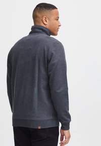 Sudadera gris con cremallera, cuello alto, mangas largas y puños acanalados. Presenta una pequeña etiqueta con el logo en el dobladillo trasero. Textura de tela suave.