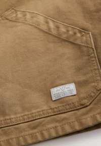 Tela de algodón marrón con un bolsillo de parche y una etiqueta blanca que dice "STANDARD ISSUE", con costuras visibles y superficie texturizada.