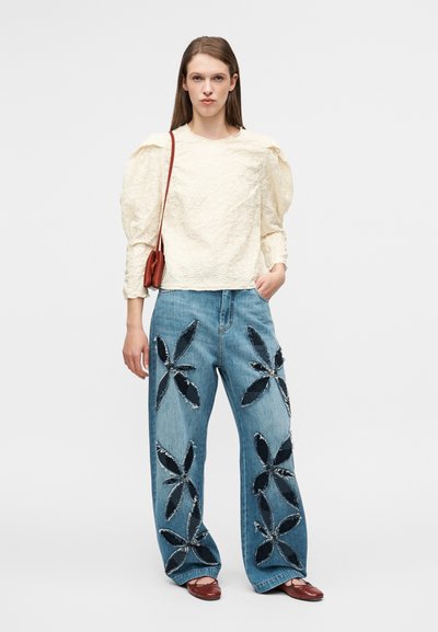 Μπλούζα με υφή κρέμας και φουσκωτά μανίκια· μπλε τζιν παντελόνι με φαρδιά μπάτσα με μαύρες λουλουδάτες εφαρμογές· το μοντέλο κρατά μία μικρή καφέ τσάντα.