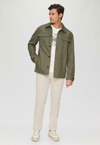 Olivengrüne Jacke mit Knopfverschluss, zwei Brusttaschen und klassischem Kragen, kombiniert mit einem weißen T-Shirt und beigen Hosen.