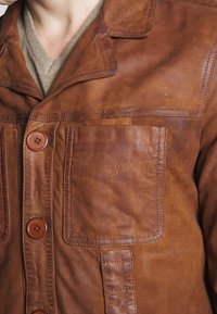 Närbild av en brun läderjacka som bärs över en beige V-ringad tröja, vilket visar knäppningar och en bröstficka fram med sömnadsdetaljer.