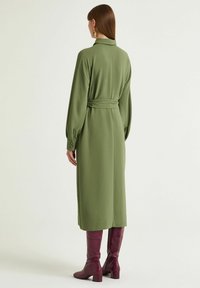 Olivgrünes, langärmliges Kleid mit tailliertem Gürtel, Kragen und geknöpften Bündchen, kombiniert mit glänzenden burgunderroten kniehohen Stiefeln.