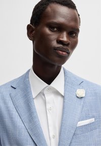 Ljusblå texturerad blazer med en vit skjorta, prydd med en dekorativ vit bröstnäsduk i form av en ros och en bröstficka med en vit näsduk.