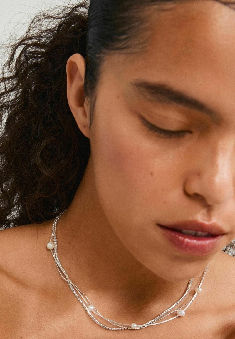 Collier en argent composé de plusieurs rangs de perles rondes, présentant un design délicat et des textures variées. Le teint de la peau est neutre.