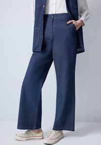 Pantalon large bleu foncé en denim, doté d'une taille haute, de poches latérales et de coutures visibles. Porté avec des baskets beiges.